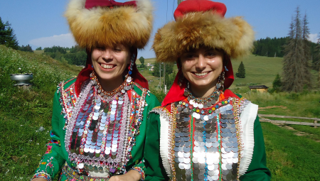 Two women wearing traditional dress, including fur hats, pose together on a hillside