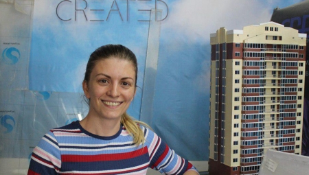 Irina, smiling in the center, stands next to a model building with her company's sign, 3D Create, in the background